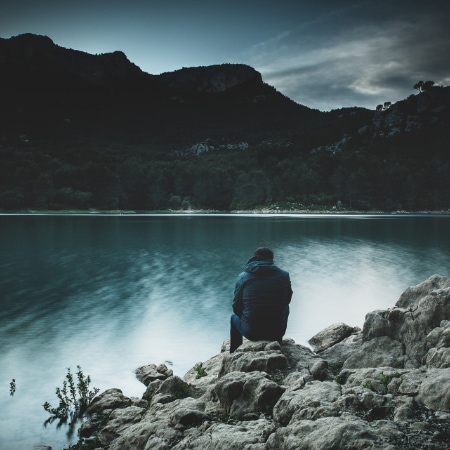 A person sits on the rocks in cold weather clothes as they face a lake and mountain landscape at dusk
