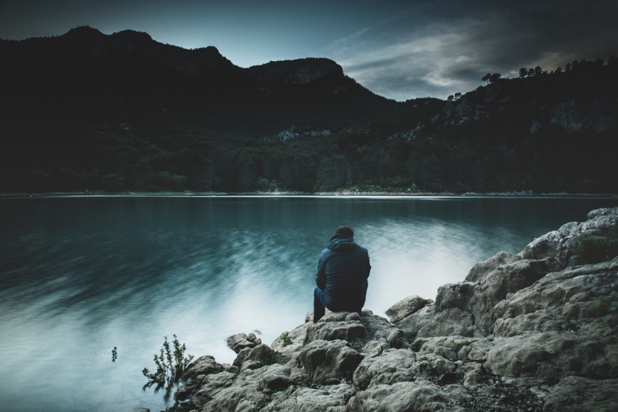 A person sits on the rocks in cold weather clothes as they face a lake and mountain landscape at dusk
