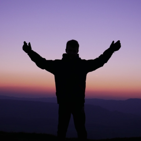 A person holds their arms up as they pray while facing toward a mountainous landscape at dusk