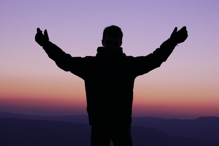 A person holds their arms up as they pray while facing toward a mountainous landscape at dusk