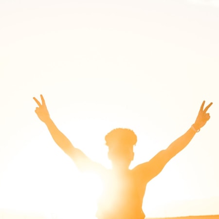 A person holding up both arms up in the air with peace signs as the sun rises behind them