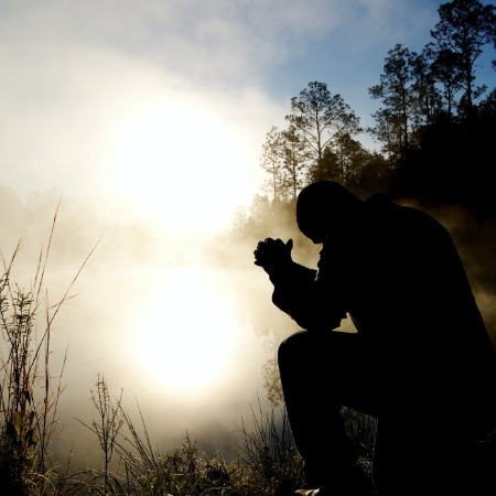 A person kneels as they pray while facing a lake in the woods at sunrise