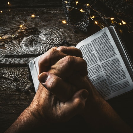 A pair of hands clasped together of an adult praying holding open a Bible on a wooden table