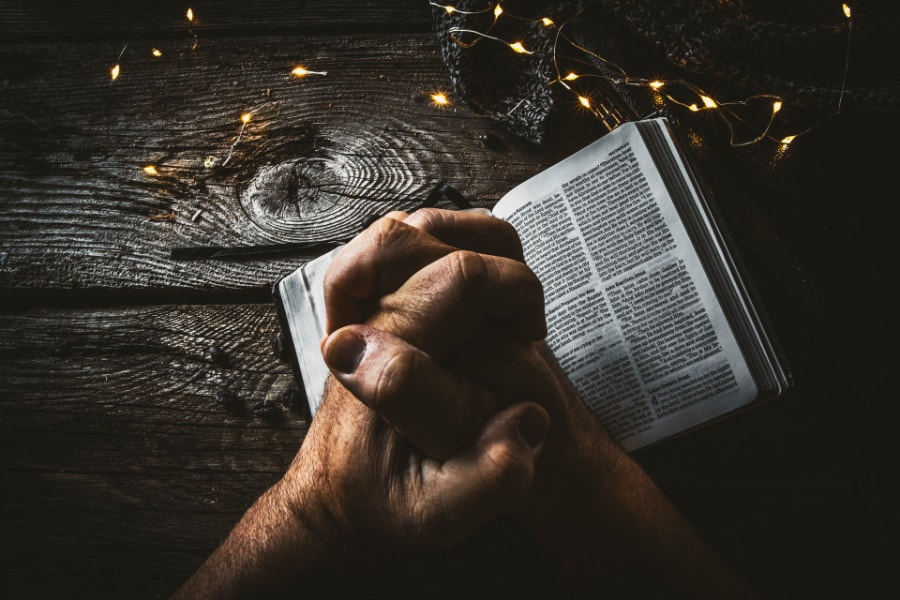 A pair of hands clasped together of an adult praying holding open a Bible on a wooden table